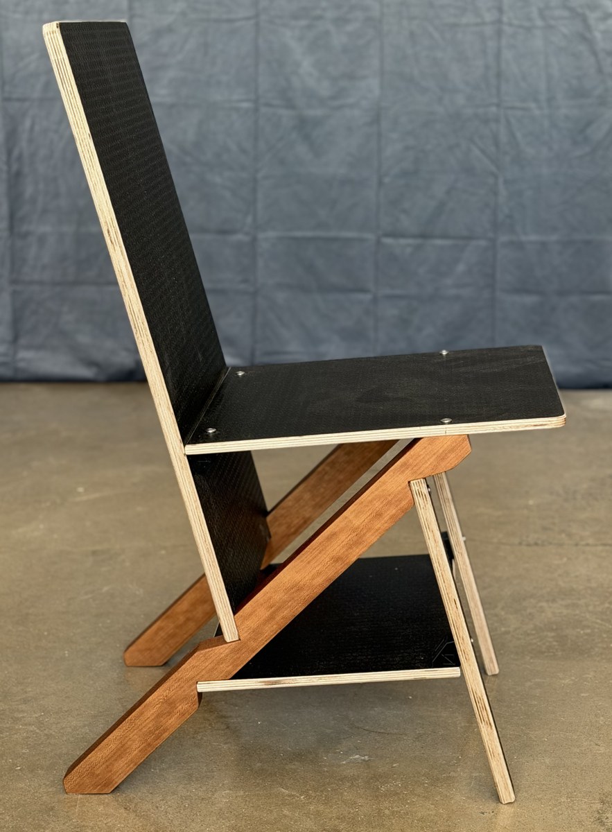 The Keel Chair in side elevation against a grey backdrop: black film-faced plywood back, seat and tie panels seen edge-on with their pale ply edges showing, a cherry keel running as one long diagonal from the top of the back down to the floor, and a pale plywood front leg crossing it